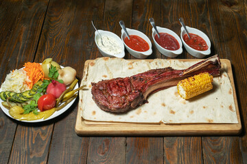 Freshly grilled Tomahawk steaks on wooden cutting board with vegetables and sauces
