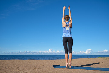 Fototapeta na wymiar Young slim girl doing yoga on the beach on a sunny morning, sun salutation stand, stretching before doing sports
