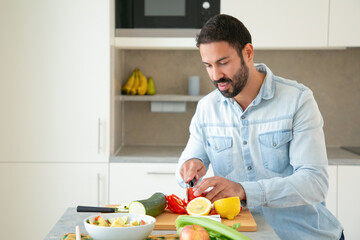 Positive handsome man cooking salad, cutting fresh vegetables on chopping board in kitchen. Medium shot, copy space. Healthy food concept