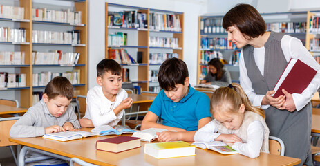 Fototapeta na wymiar Group of cheerful school kids studying in school library with friendly female teacher