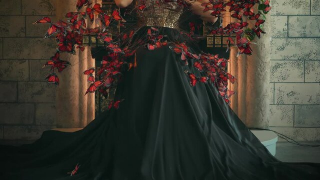 Magical fantastic woman sitting vintage bronze armchair. Beauty luxurious elegant outfit long black silk skirt, sparkling chic bodice. Girl princess enjoy beautiful creative artificial red butterflies