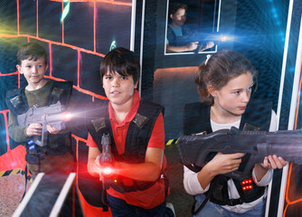 Positive teen girl and boys with laser guns playing laser tag on dark labyrinth