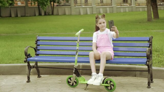A cute beautiful girl sits on a bench in the park and takes a selfie.