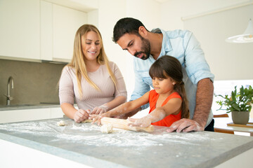 Fototapeta premium Positive parents watching daughter rolling dough on kitchen desk with flour messy. Young couple and their girl baking buns or pies together. Family cooking concept