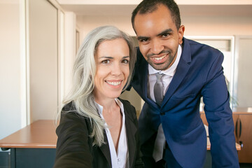 Cheerful professional partners posing for photo, smiling and looking at camera. African American successful office employer and Caucasian businesswoman taking selfie. Teamwork and business concept
