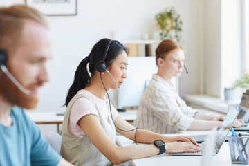 Fototapeta premium Asian young businesswoman in headset typing on laptop at her workplace with other colleagues working at office