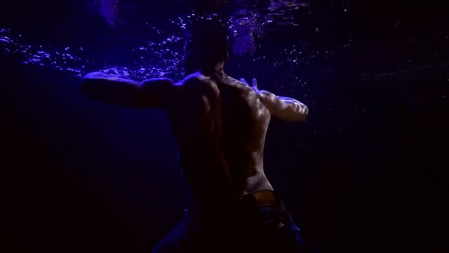 sexy man with naked muscular torso is moving underwater in darkness, dancing by hands