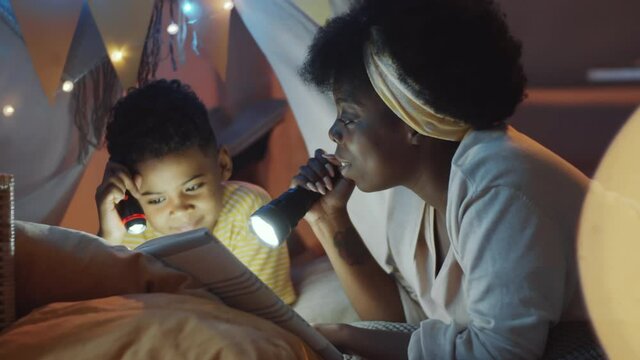 Cheerful African American mother and little son lying in teepee tent decorated with lights, holding flashlights and reading fairytale story aloud while spending evening together at home