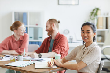 Obraz premium Portrait of Asian young woman smiling at camera while sitting at the table with other business people at meeting at office