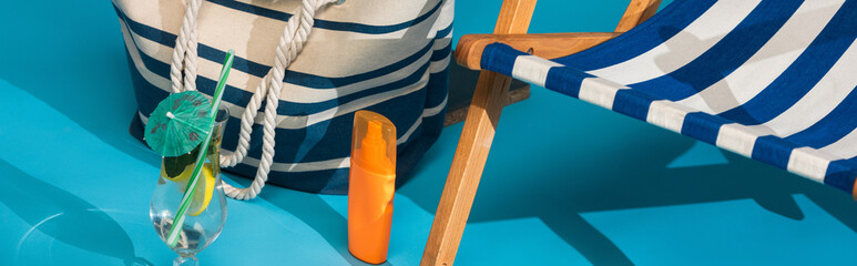 close up view of striped deck chair near sunscreen, beach bag and cocktail on blue background,...