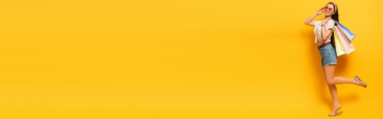 full length view of stylish summer brunette girl with shopping bags on yellow background, panoramic shot