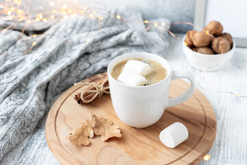 White mug with coffee and marshmallow, knitted scarf and garland on wooden table. Autumn mood. Cozy autumn composition. Hygge concept
