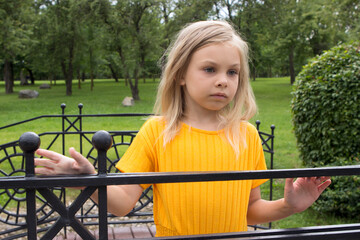 A little girl of six-seven years old with long blond hair in a yellow dress suit stands in a summer park on a decorative bridge.