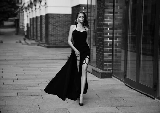 Film grain added. Sexy girl in classic evening trendy dress. Black and white photo. Luxury vintage woman with long leg. Beautiful retro Lady. Glamour queen. Noir fashion model walks down old street 