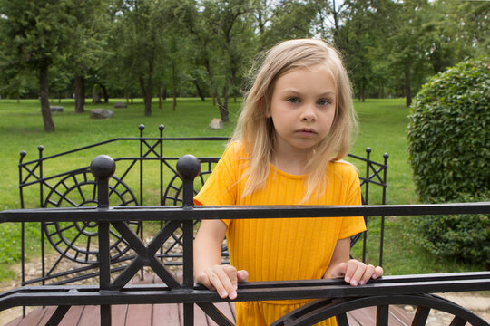 A little girl of six-seven years old with long blond hair in a plain yellow outfit stands and smiles in a summer park on a decorative bridge and poses.