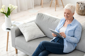 Modern senior woman relaxing on couch at home with digital pad