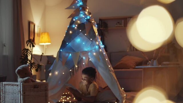 Tilt down shot of adorable little African American boy lying in cozy teepee decorated with lights, smiling and playing on digital tablet in dark room in the evening