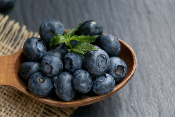 Several blueberries and green mint leaves on a wooden spoon and a burlap napkin. Black stone background. Healthy snack.