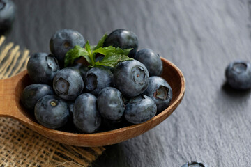 Several blueberries and green mint leaves on a wooden spoon and a burlap napkin. Black stone background. Healthy snack.