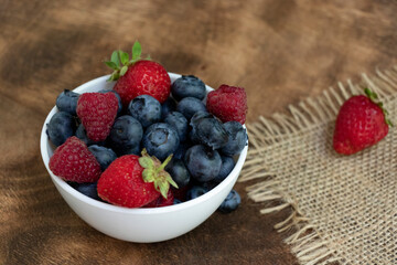 Set of summer berries in a small white bowl on a napkin and wooden background. Blueberries, raspberries, and strawberries are a varied healthy snack. Yummy.