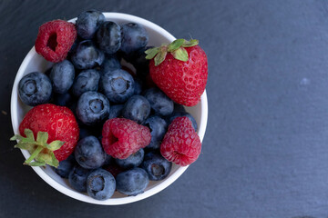 Set of summer berries in a small white bowl on a blackboard. Blueberries, raspberries, and strawberries are a varied healthy snack. Yummy.