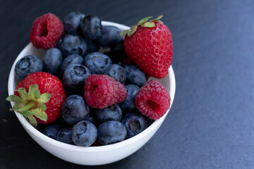 Set of summer berries in a small white bowl on a blackboard. Blueberries, raspberries, and strawberries are a varied healthy snack. Yummy.