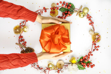 Gold gift box with big red bow in hands on white background with christmas decoration around.