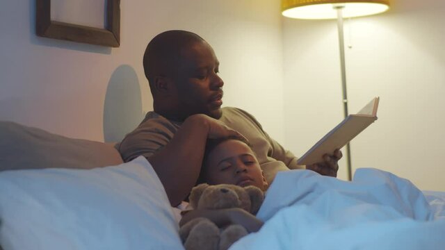 Loving African American dad reading fairytale and caressing cute little son while he sleeping with toy under blanket on bed at night