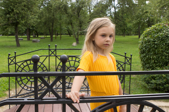 A little girl of six-seven years old with long blond hair in a yellow dress suit stands in a summer park on a decorative bridge.