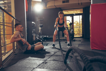 Fototapeta na wymiar Beautiful young sporty couple workout in gym together. Caucasian man training with female trainer. Concept of sport, activity, healthy lifestyle, strength and power. Working out with ropes.