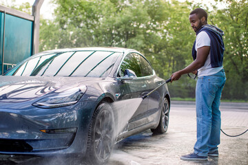 Handsome African guy in t-shirt and jeans washing his blue modern electric luxury car, rinsing the...