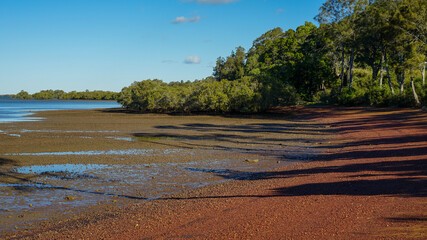 Shadows on the red pebbly beach at low tide, with trees, mangroves and blue sea. Redland Bay, Queensland, Australia.	
