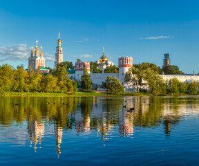 Obraz premium The famous Moscow landmark with Golden domes and a reflection of the landscape on the water of the Park pond against the background of a picturesque blue sky with clouds.