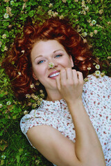 portrait beauté romantique et nature d'une jeune et jolie femme rousse souriante aux cheveux bouclés dans un parterre d'herbes vertes et pâquerettes en été 