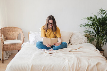 Young beautiful girl student in glasses, an orange T-shirt and blue jeans sits on bed with pillows and reads paper book. Self-education concept at home during quarantine. Home schooling, hobby.