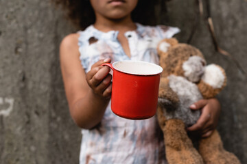 Cropped view of poor african american kid holding metal cup and teddy bear while begging alms on...