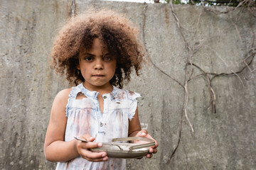 Selective focus of african american kid with messy face holding metal plate and spoon on urban...