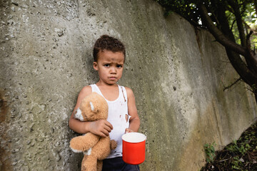 Displeased african american boy holding dirty teddy bear and metal cup near concrete wall on urban...