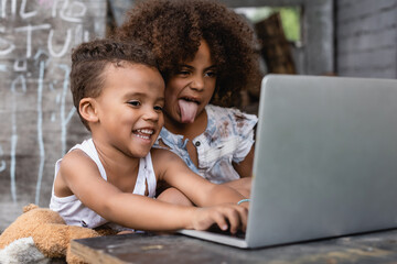 selective focus of poor african american kid sticking out tongue near happy kid using laptop outside