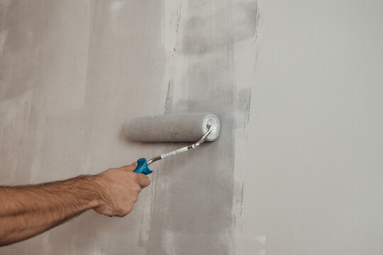 Male hand holding industrial paint roller. Man painting white wall with grey paint.