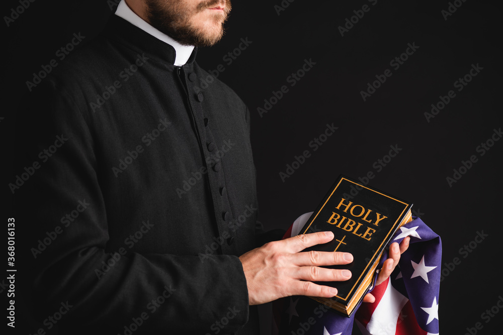 Wall mural partial view of priest holding holy bible and american flag isolated on black - Wall murals