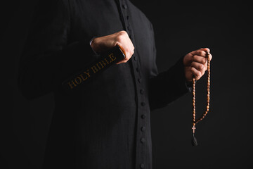 Cropped view of pastor holding holy bible and rosary beads in hands isolated on black