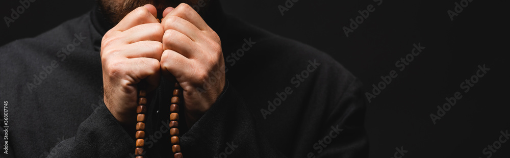 Wall mural panoramic crop of priest holding rosary beads in hands isolated on black - Wall murals