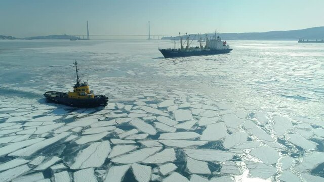 Picturesque view of Yellow tugboat, cargo ships vessel sailing in winter frozen icy Bosphorus strait Vladivostok unique cityscape. Cracked ice pattern in sea. Russian bridge. Sunny day Aerial sideways