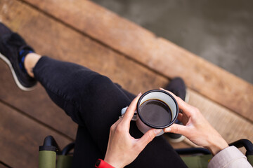 A girl holds a metal cup of coffee in her hands near the lake while sitting on a wooden pier. Close-up, top view.