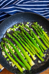 Tasty green asparagus is fried in a pan with garlic, next to it is a blue striped towel. Close-up.