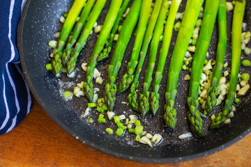 Tasty green asparagus is fried in a pan with garlic, next to it is a blue striped towel. Close-up.