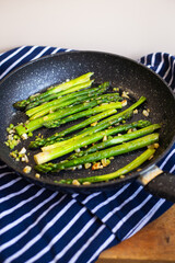 Tasty green asparagus is fried in a pan with garlic, next to it is a blue striped towel. Close-up, top view.