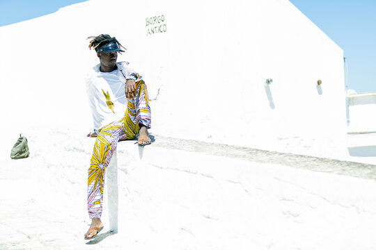 Italian immigrant man of sub-Saharan origin with sun hat and black and golden hair with an elegant look with the traditional colors of Ghana sitting on the wall of the belvedere of the white city