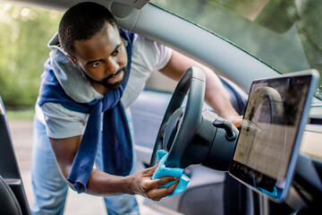 Handsome African young man in casual wear cleaning car interior, car steering wheel using microfiber clothes. Car detailing or valeting concept. Selective focus